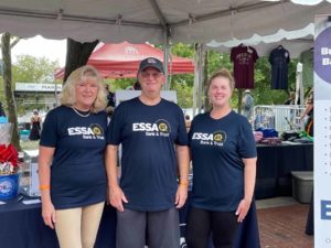 3 ESSA employees pose near a table of ESSA swag and brochures doing a concert.