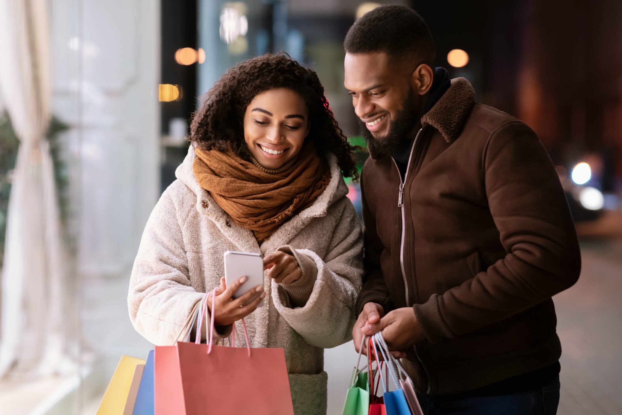 Choosing Festive Present. Smiling african american lady showing her smartphone to boyfriend or husband, pointing at screen, guy holding shopping bags, standing outside on the street. Discount And Sale