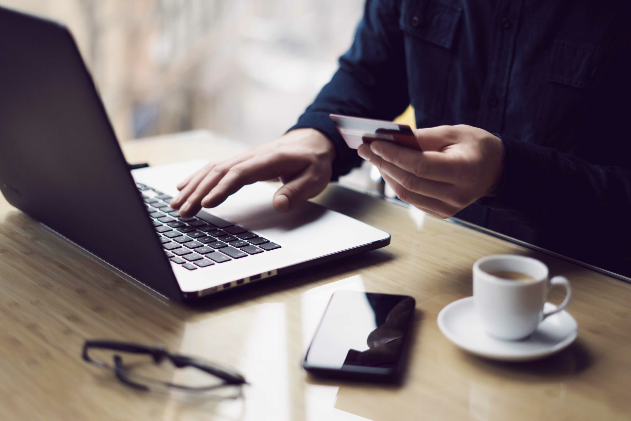 Businessman holding plastic credit card in hand and using laptop computer while sitting at the wooden table.Man making online money transfer.Blurred background.Horizontal